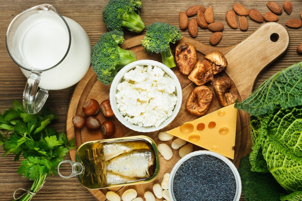 Top-down view of a wooden board showcasing a variety of calcium-rich foods including assorted cheeses, walnuts, broccoli florets, canned sardines, and a glass jug of fresh milk.