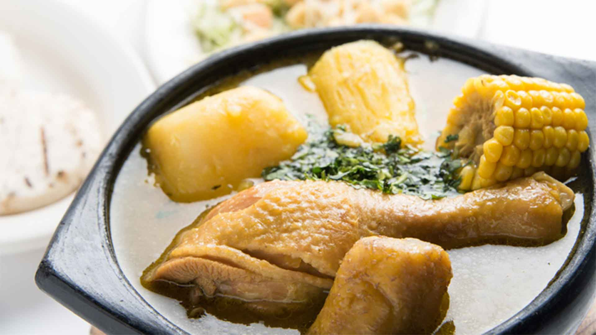 A hearty bowl of Colombian Sancocho, a comforting soup filled with chicken, corn on the cob, plantain, beans, and yuca, served with a side of rice, avocado, and ají dipping sauce for a customizable and satisfying meal.