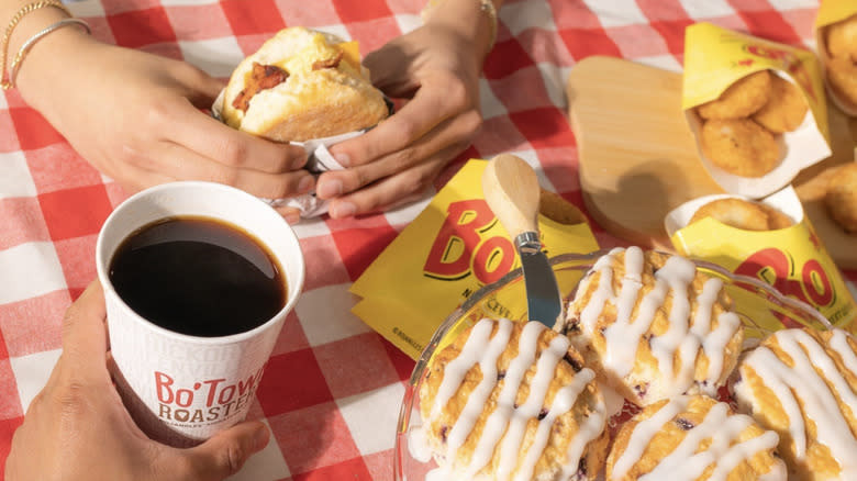 Hands holding a biscuit sandwich and a coffee with biscuits and hash browns on the table