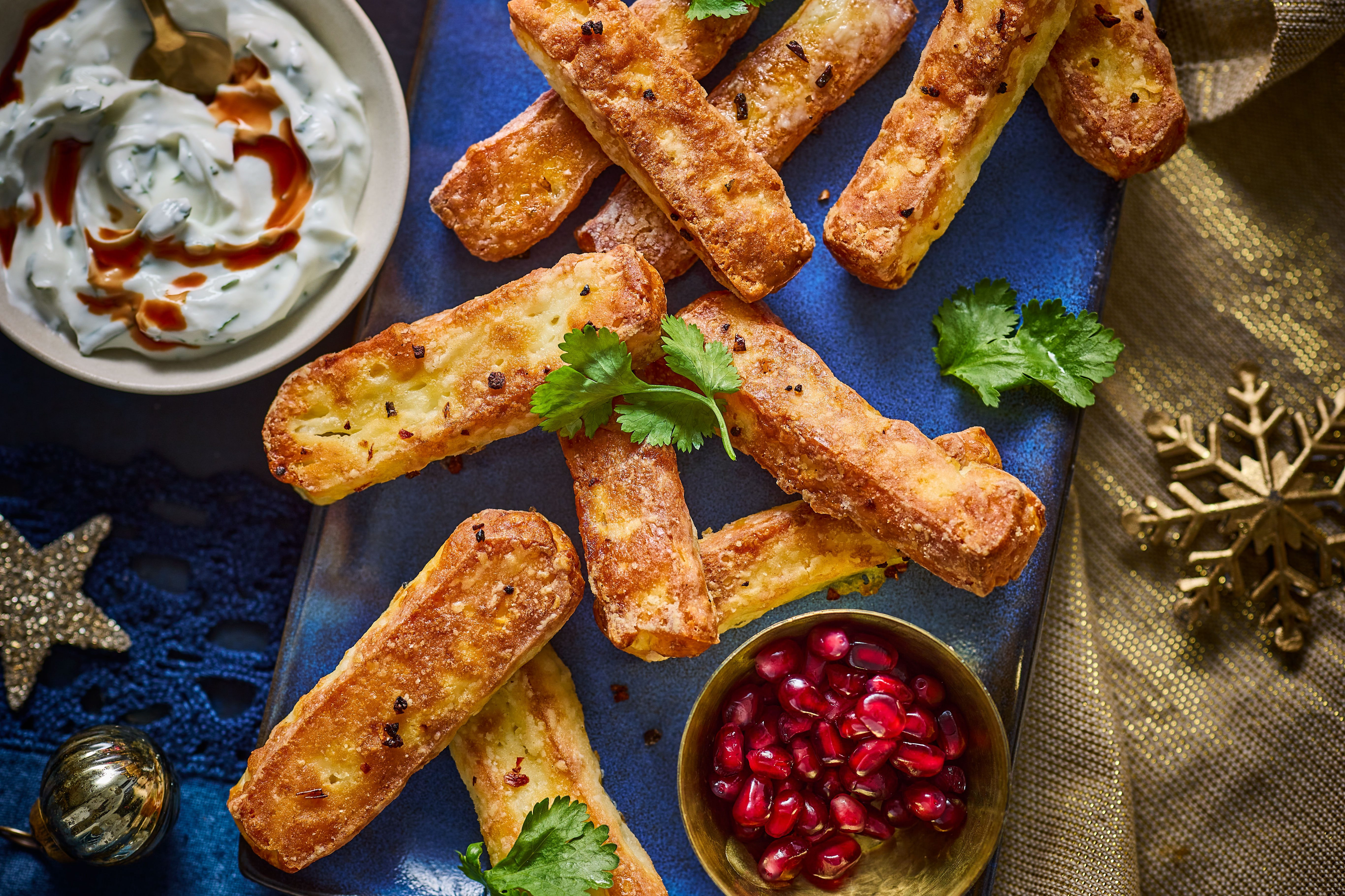 Halloumi sticks on a blue board next to a bowl of pomegranates and a bowl of yogurt