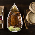 A festive table setting with various Chinese dishes, including dumplings, spring rolls, and noodles, decorated with Christmas ornaments and lights, symbolizing the fusion of cultures during the holiday season.