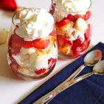 Layers of fresh strawberries, toasted angel food cake and whipped cream in clear glasses with spoons next to the glasses